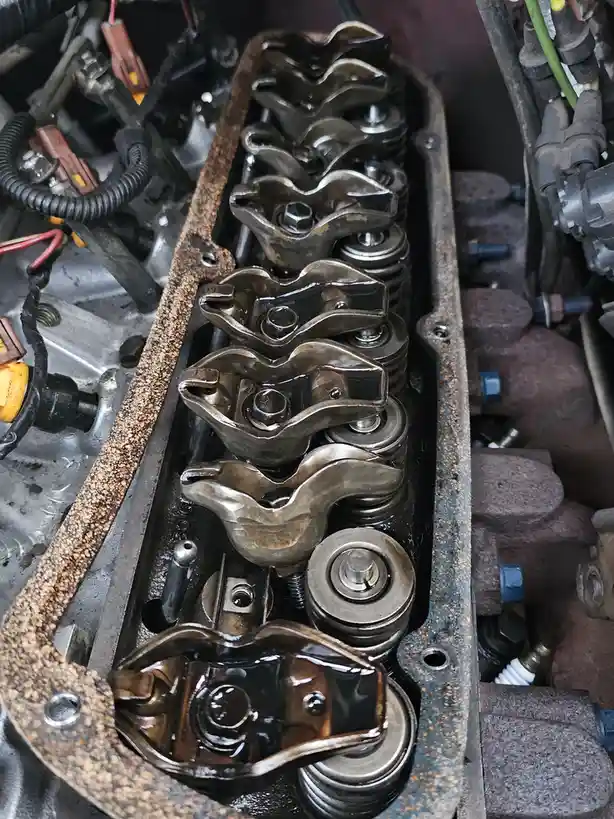 A close-up photo of a car engine's valve train with visible rocker arms, springs, and oil residue, surrounded by engine…