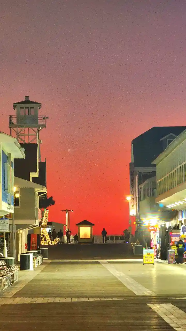 A vibrant sunset with a gradient of pink and red skies over a boardwalk lined with shops and a water tower. Silhouettes of…