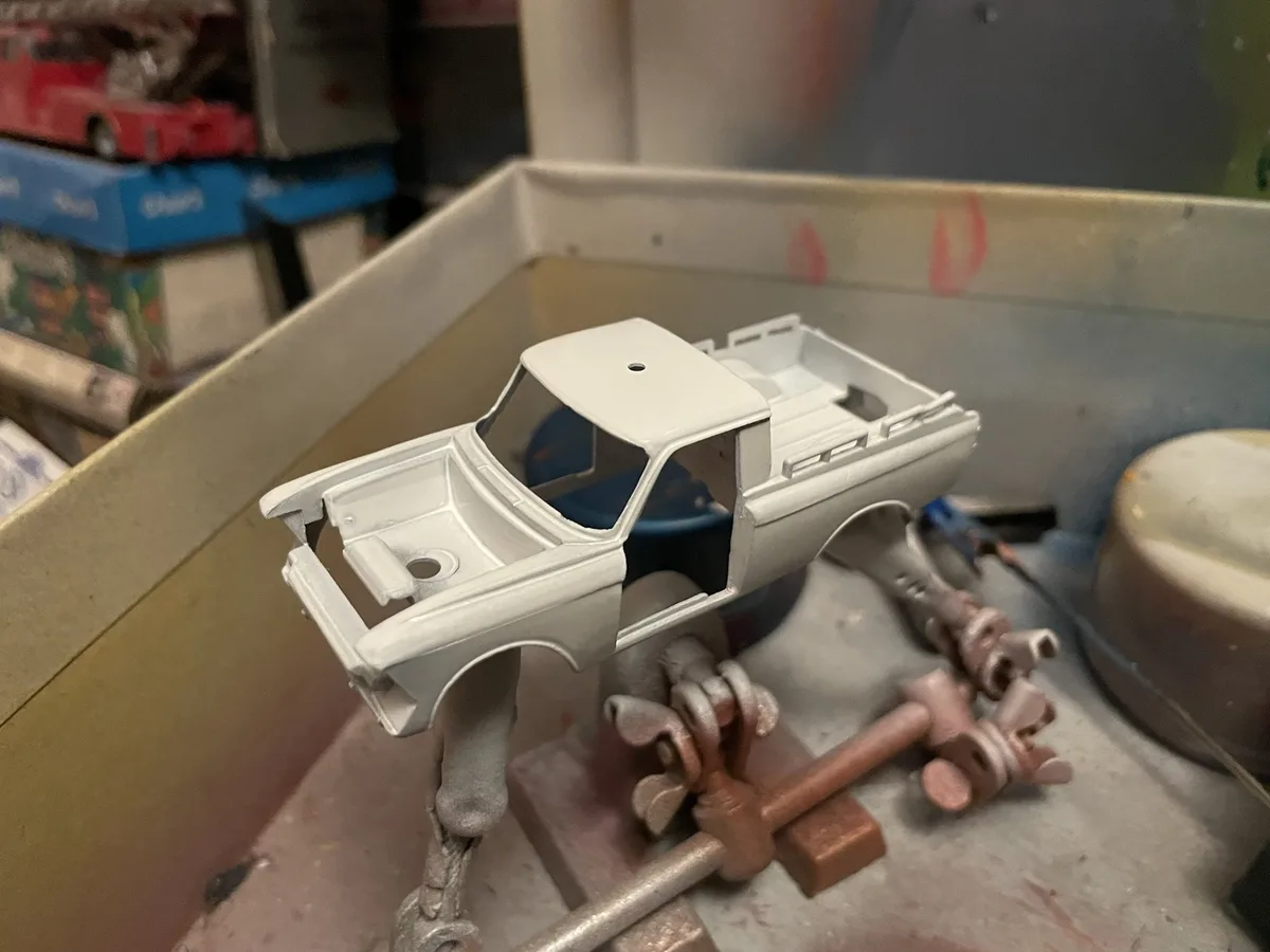 A white-painted, unpainted plastic model truck body sits on a workbench, surrounded by tools and parts, with the overlaid…