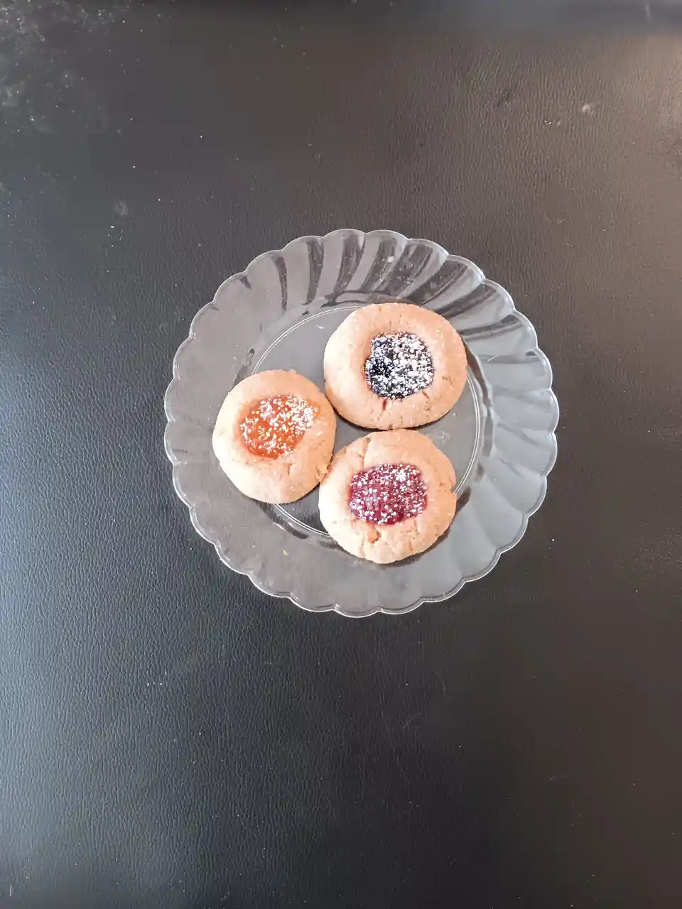 Three thumbprint cookies with different colored jam centers (orange, dark purple, and red) sit on a clear glass plate with a…