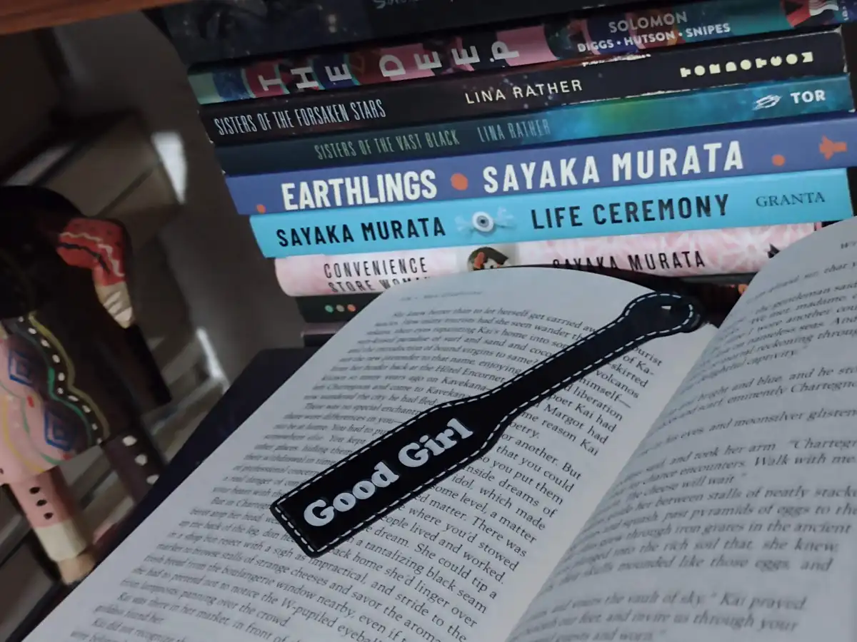 An open book with a black bookmark that reads 'Good Girl' in white text lies on a stack of books including titles by Sayaka…