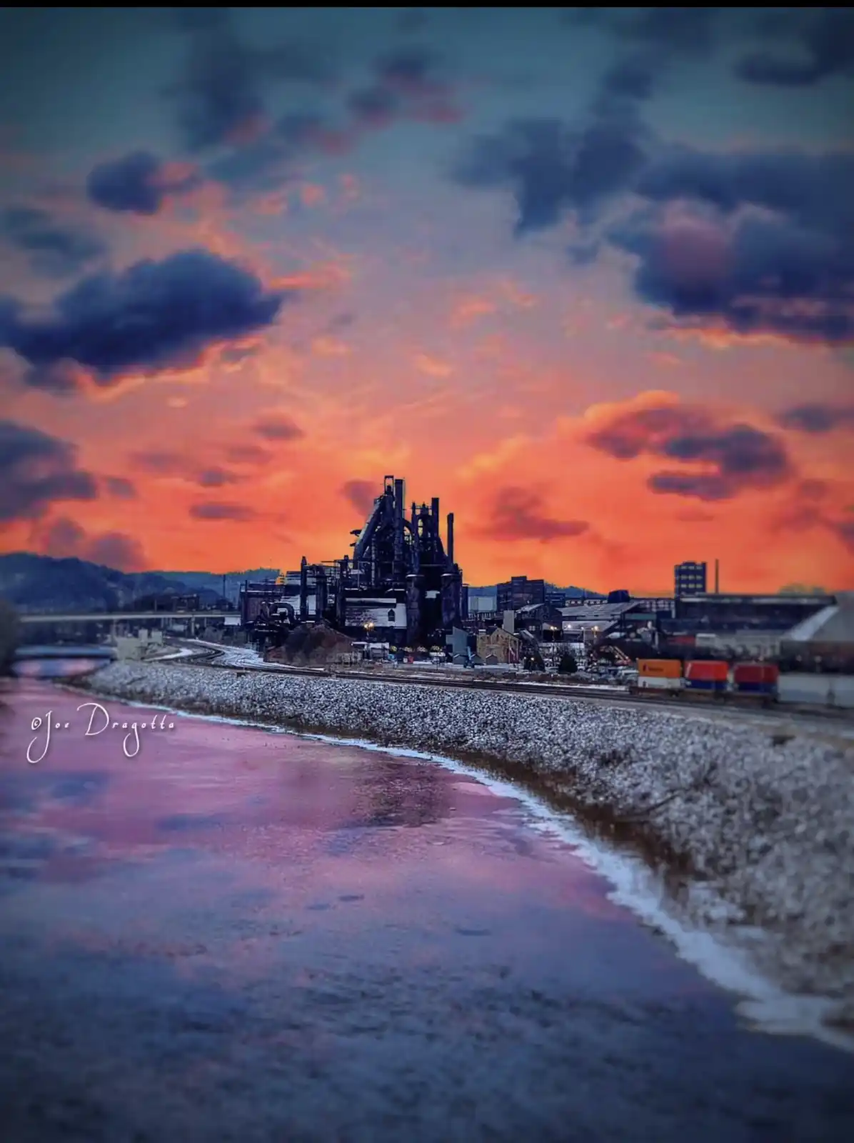 A dramatic photograph shows a large, dark industrial steel mill complex at sunset, with vibrant orange and purple clouds…