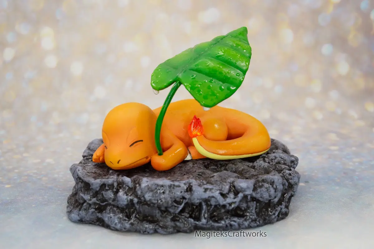 A small orange figurine of the Pokémon Charmander is curled up sleeping on a dark grey rock base. A large green leaf is…