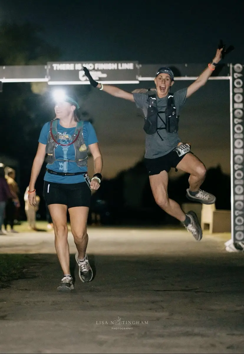 Two female runners at night under a banner reading 'THERE IS NO FINISH LINE'. One woman in a blue shirt and headlamp walks…