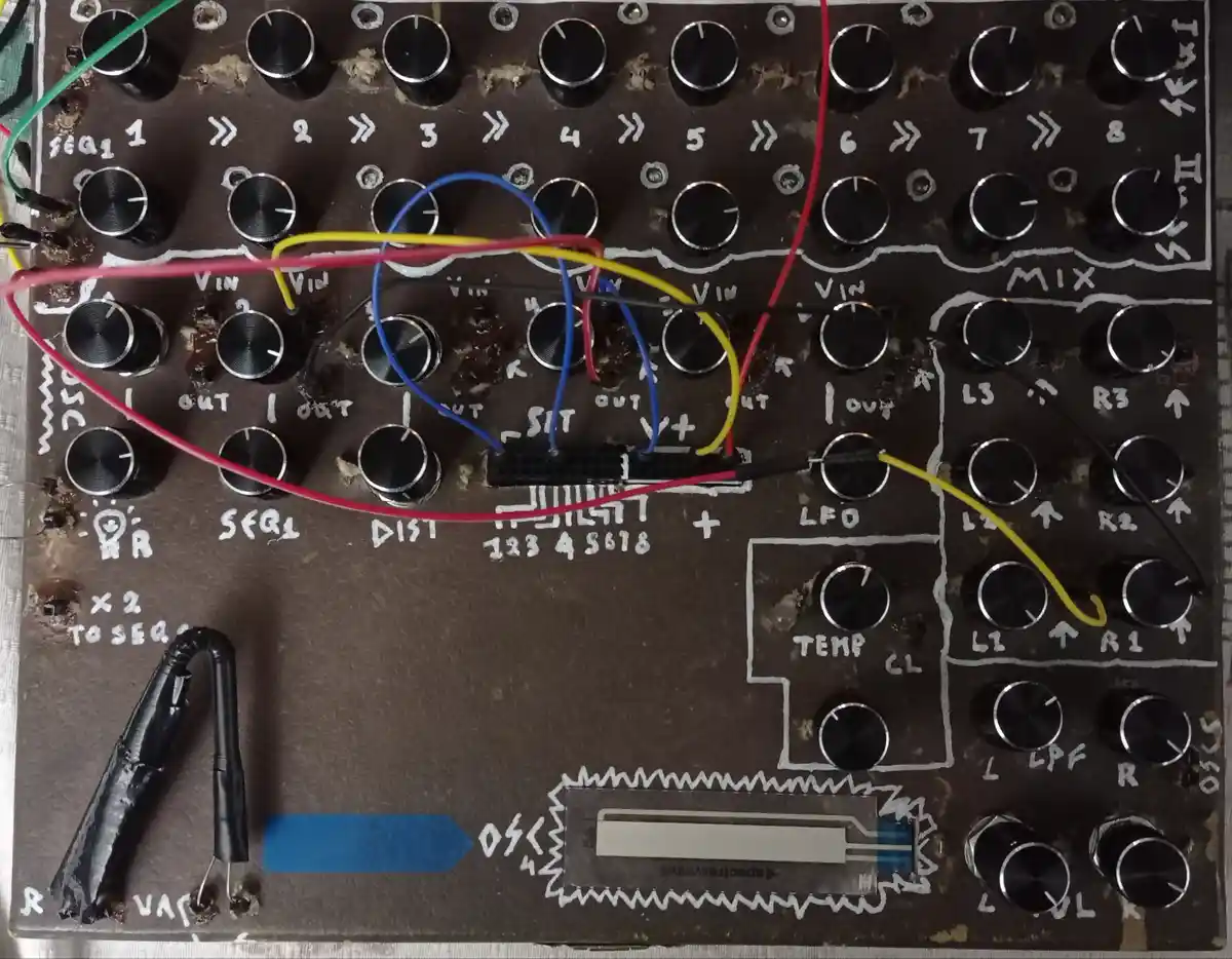 A close-up of a homemade, DIY electronic music synthesizer module with numerous black knobs, hand-drawn white labels like…