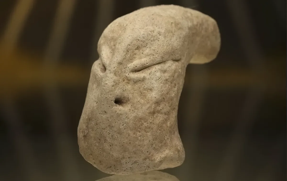 a close-up photograph of a rough, beige stone sculpture shaped vaguely like a human face, featuring a single closed eye and…