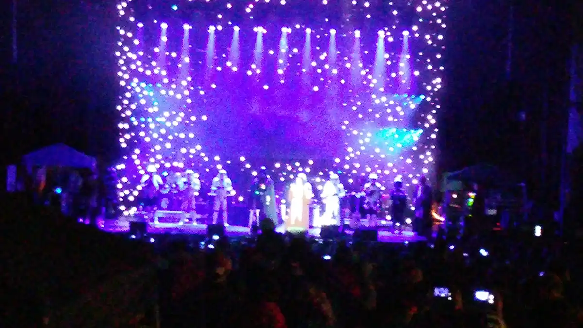 A blurry photo of a concert stage bathed in purple and blue lights, with performers silhouetted against a backdrop of bright…