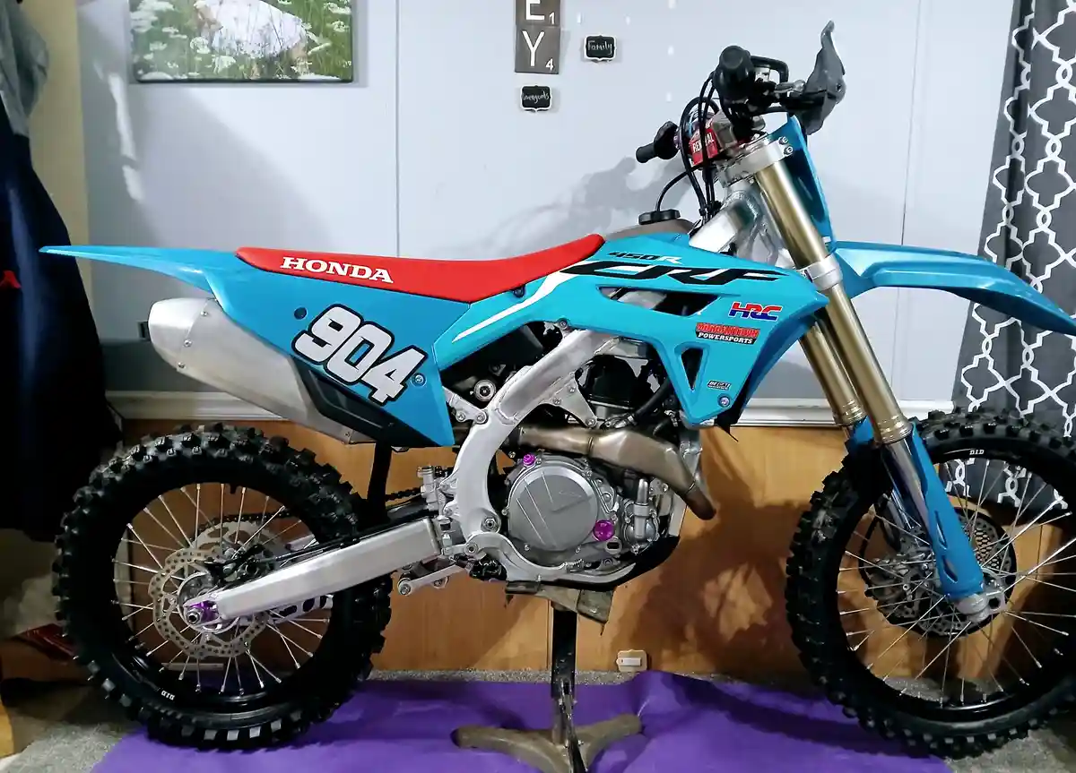 A blue and red Honda dirt bike with the number 904 prominently displayed on its side is parked indoors on a purple mat. The…
