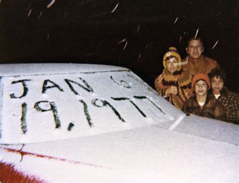 A snowy, nighttime photo shows three people posing behind a white car whose hood is covered in snow. The snow has been…