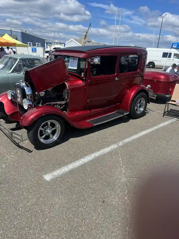 A shiny red vintage car with its hood open is parked at an outdoor car show under a partly cloudy sky. The car has chrome…