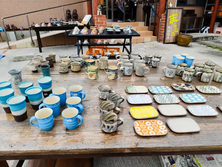 A wooden table outdoors displays dozens of handmade ceramic mugs and square plates in various colors and patterns, with a…