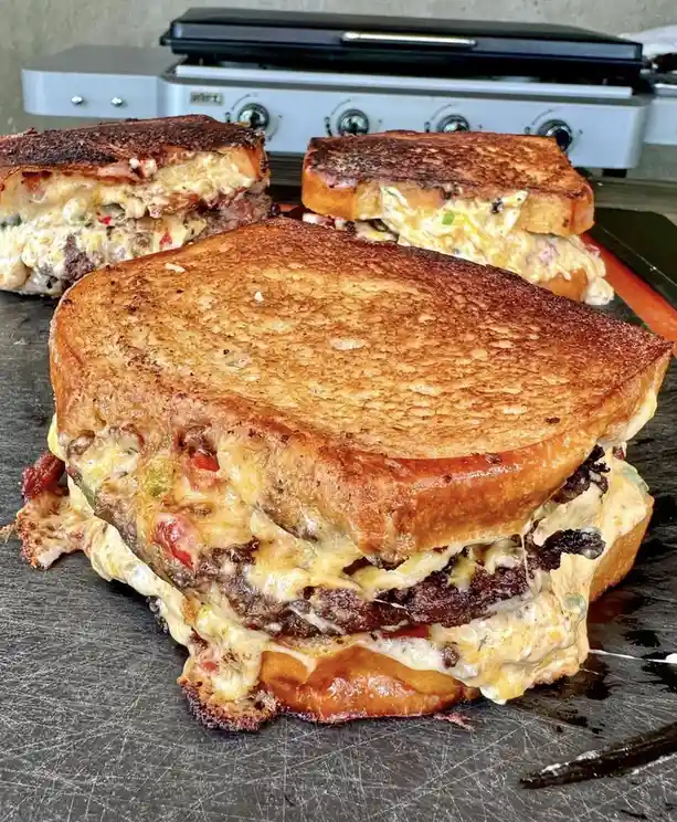 Three large, golden-brown grilled sandwiches with melted cheese and meat peeking out are displayed on a metal surface, with…