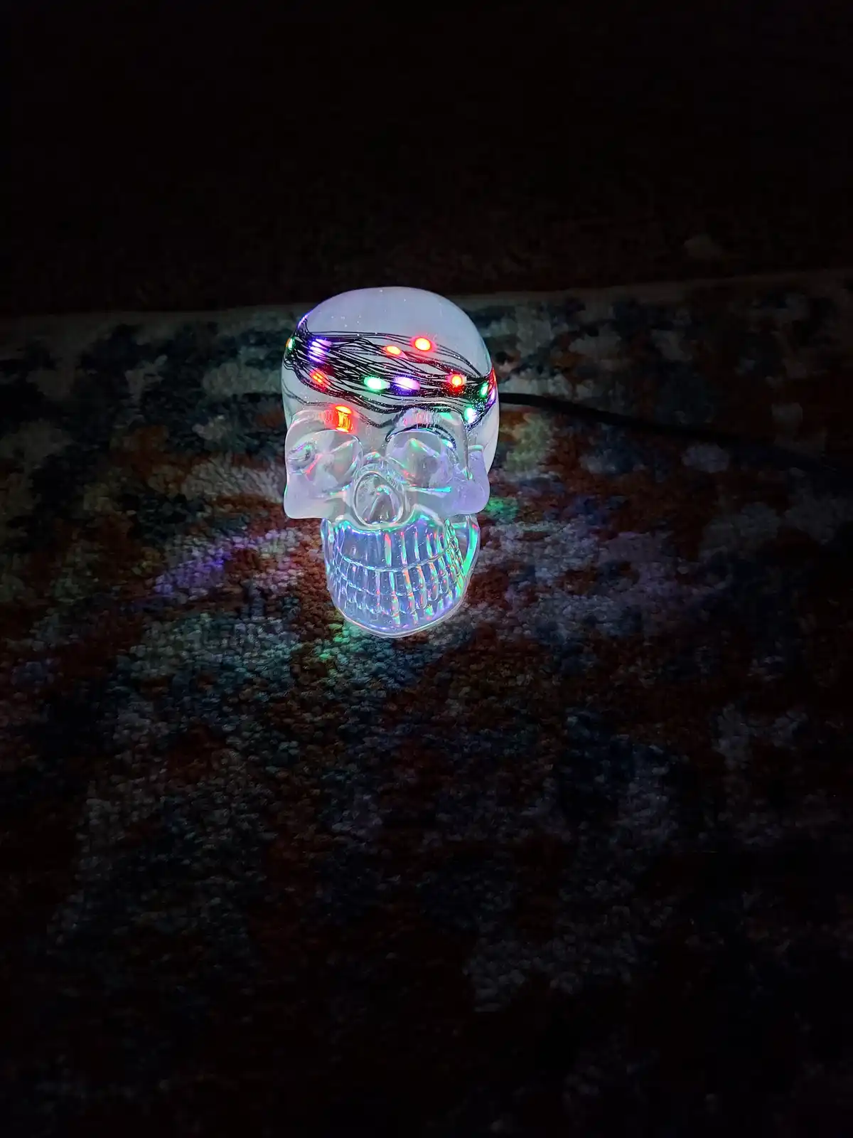 A clear plastic skull decoration with embedded multicolored LED lights is sitting on a dark patterned carpet in a dimly lit…
