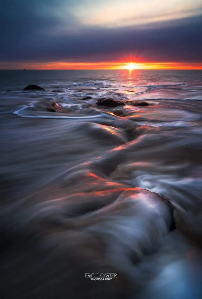A long-exposure photograph of a sunset over the ocean, where the water appears smooth and flowing around rocks, with vibrant…