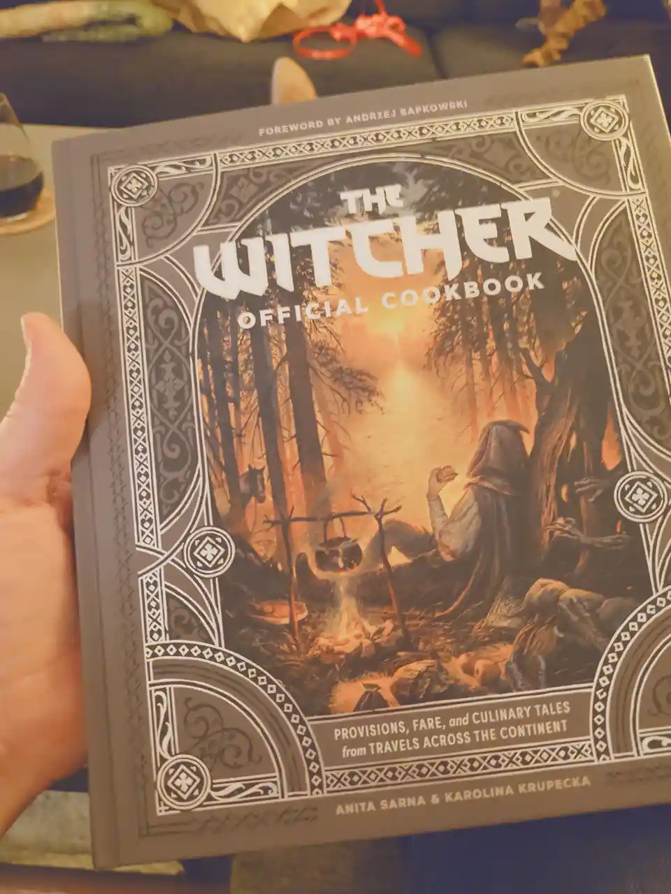 A hand holds up 'The Witcher Official Cookbook' featuring a fantasy illustration of a hooded figure cooking over a campfire…