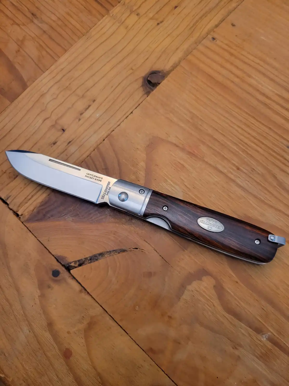 A Fällkniven brand folding pocket knife with a wooden handle and stainless steel blade lies open on a light brown wooden…