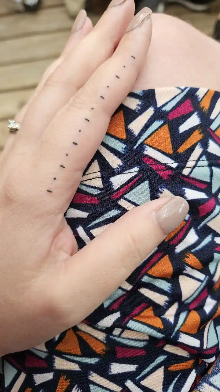 A close-up photo shows a person's hand resting on their knee, with a line of small black dots tattooed along the side of…