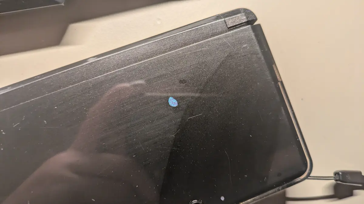 A close-up of a black, heavily scratched laptop lid with a small, iridescent blue sticker near the hinge. The surface…
