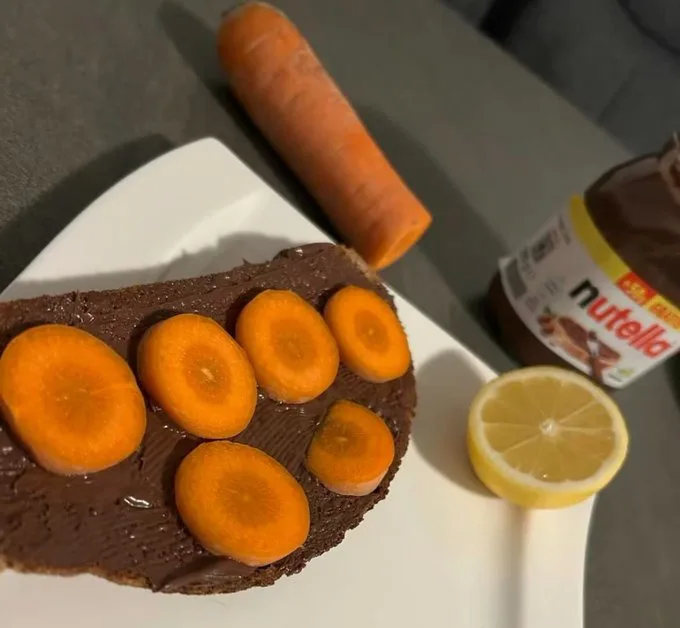 A slice of bread covered in Nutella is topped with sliced carrots arranged like eyes and a mouth, next to a whole carrot, a…