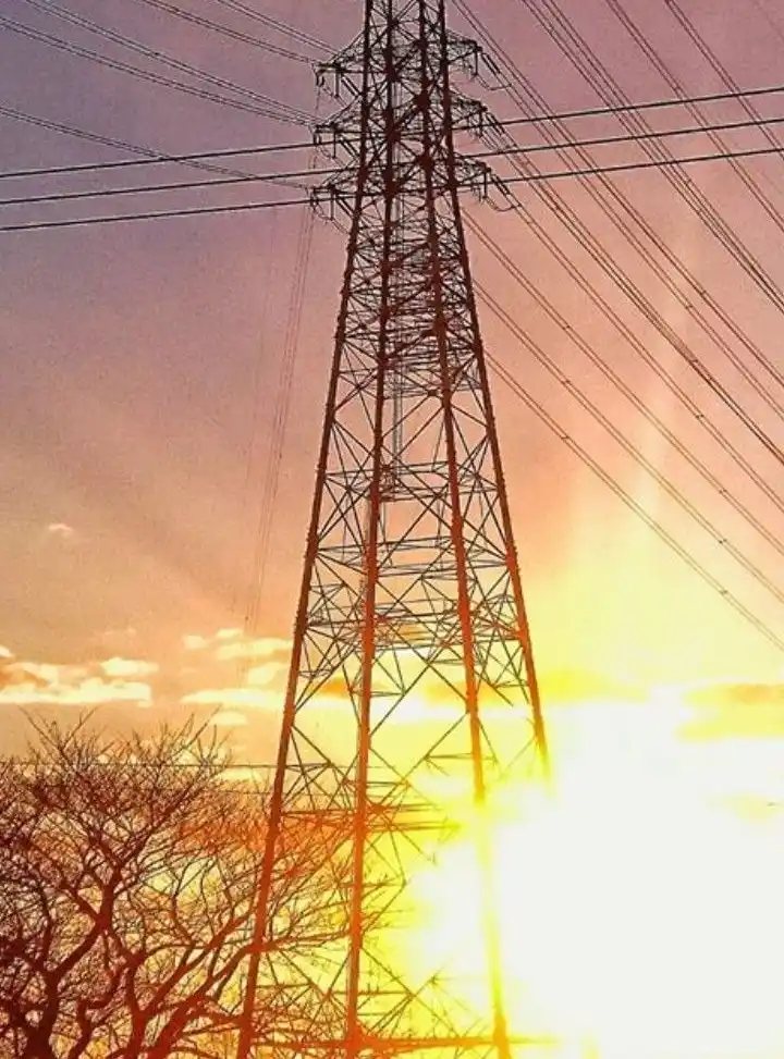 A tall metal electricity pylon stands against a vibrant sunset sky, with the sun's bright glow behind it and bare tree…