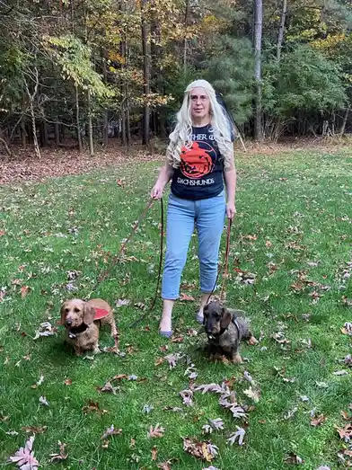 A person with long blonde hair and glasses stands in a grassy yard with fallen leaves, holding leashes for two dachshunds…