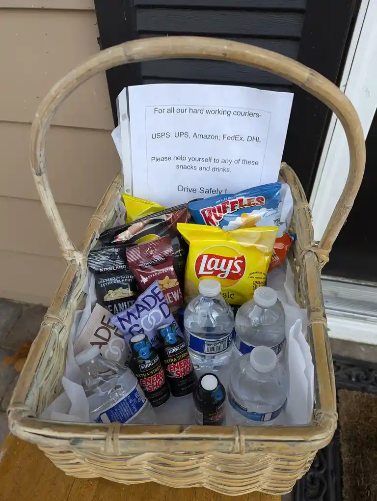 A wicker basket filled with snacks and drinks sits on a porch, including Lay's chips, Ruffles, energy shots, and water…