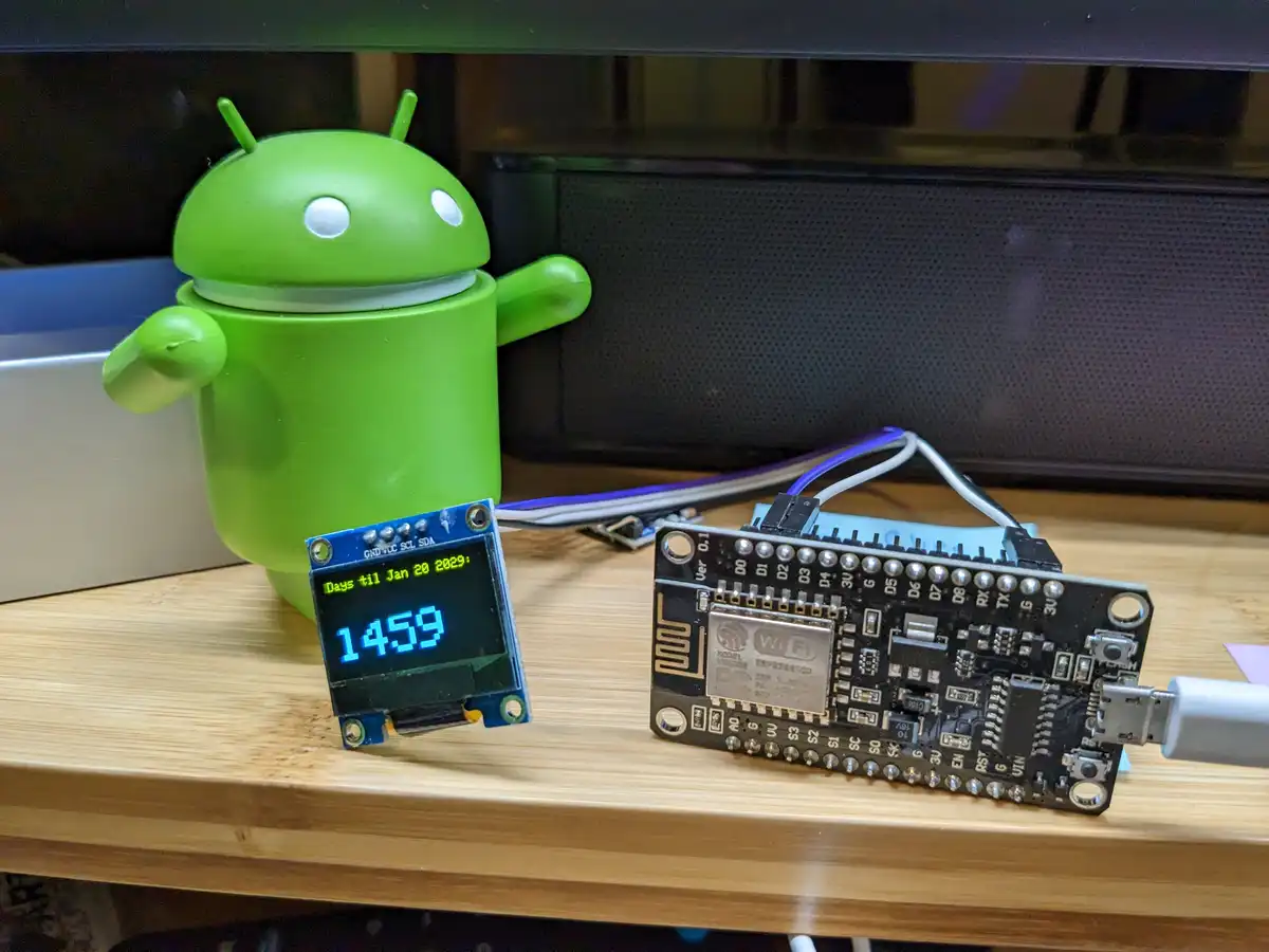 A green Android mascot figurine stands beside an electronic circuit board and a small OLED display showing 'Days til Jan 20…