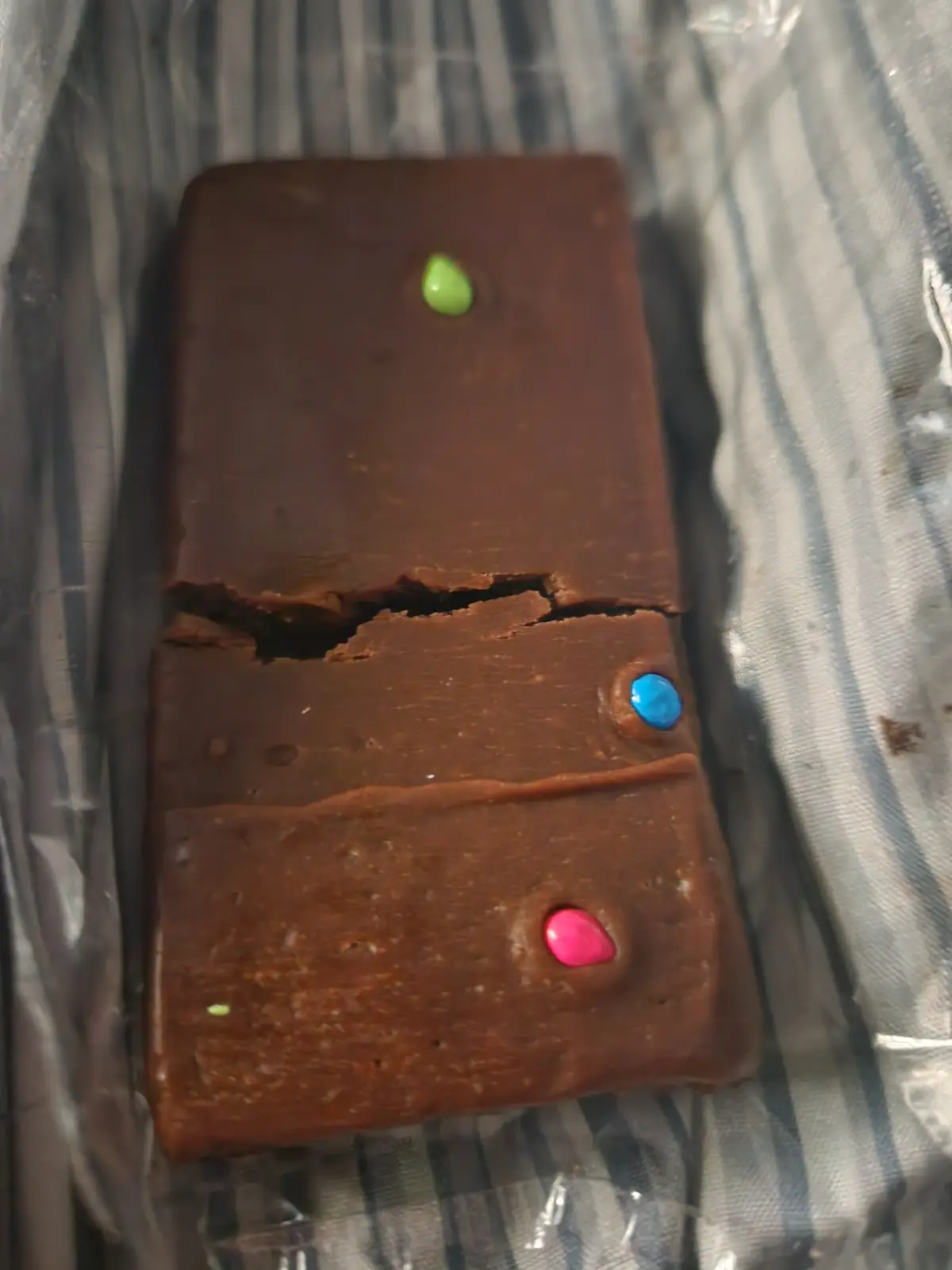 A broken chocolate bar with three colorful candy pieces embedded in it, resembling a cracked smartphone with buttons, lies…