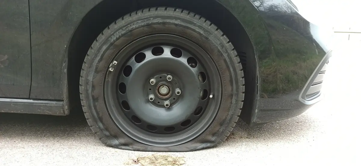 A close-up photo shows a flat, deflated car tire on a dark-colored vehicle, with the wheel rim and lug nuts visible. The…