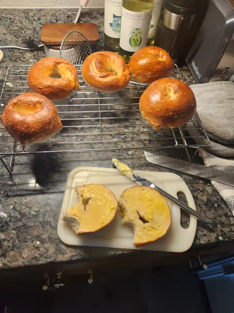 A kitchen counter displays five golden-brown bagels cooling on a wire rack, while one bagel is sliced open on a white…