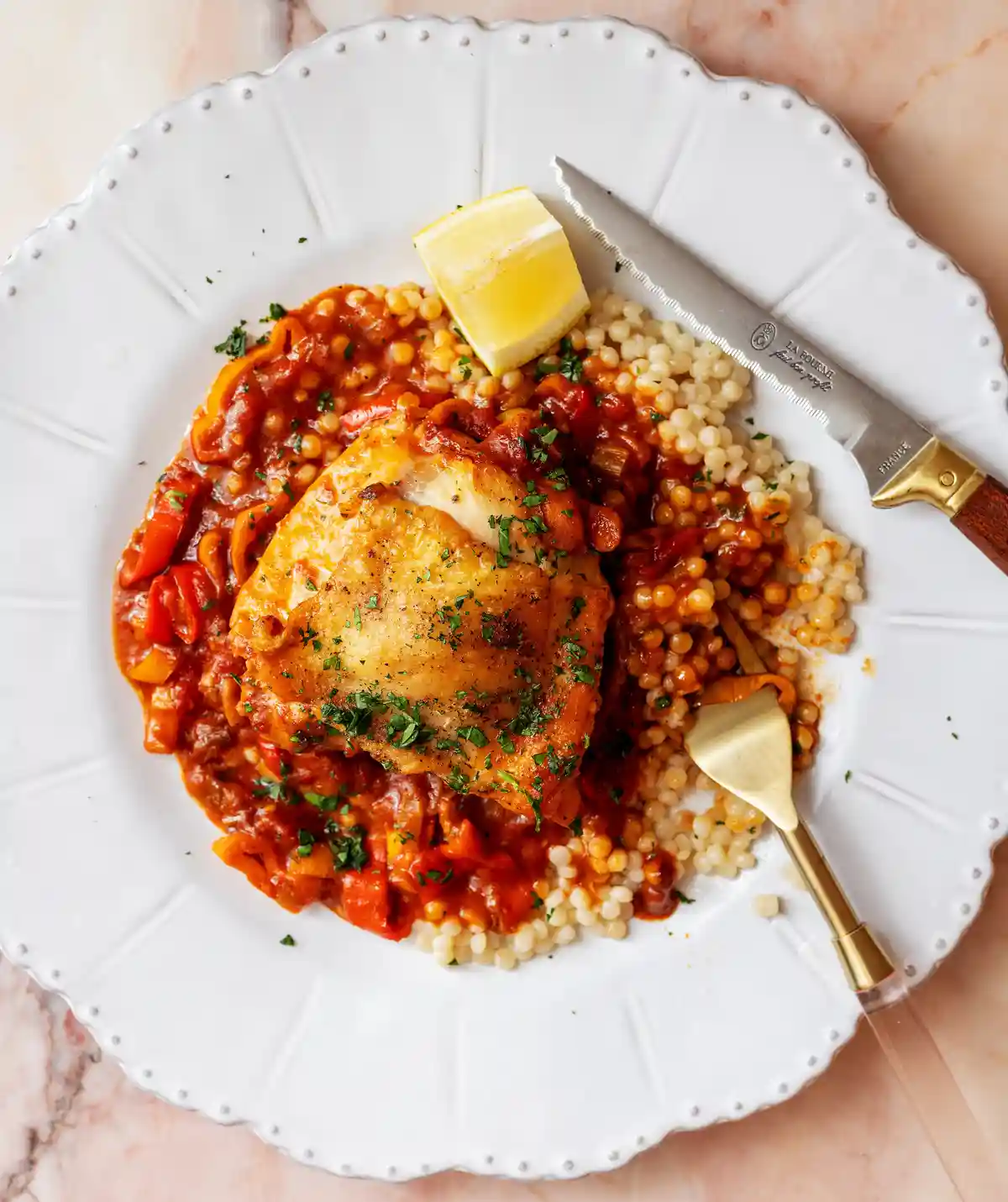 A professionally styled overhead photo of a white plate holding a dish of chicken thigh in a red tomato sauce over couscous…
