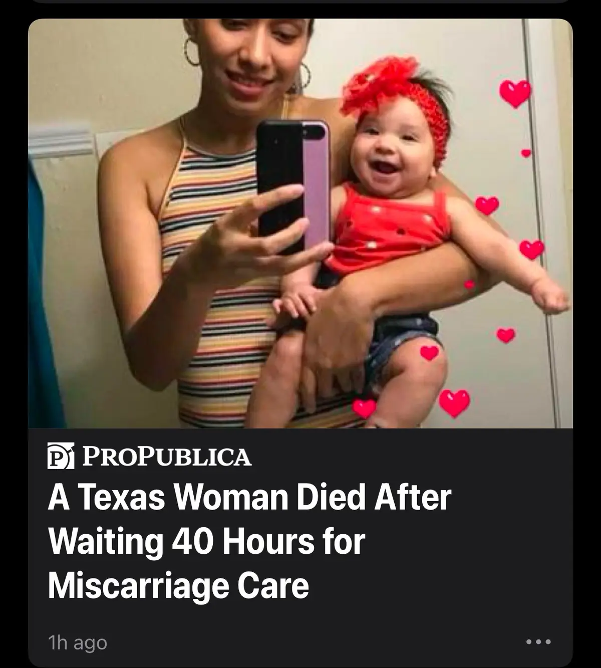 A woman in a striped tank top holds a smiling baby wearing a red outfit and headband while taking a selfie in a mirror. Red…