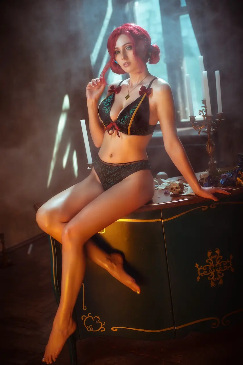 A woman with red hair styled in buns sits provocatively on a dark green ornate cabinet in a dim, atmospheric room with smoke…