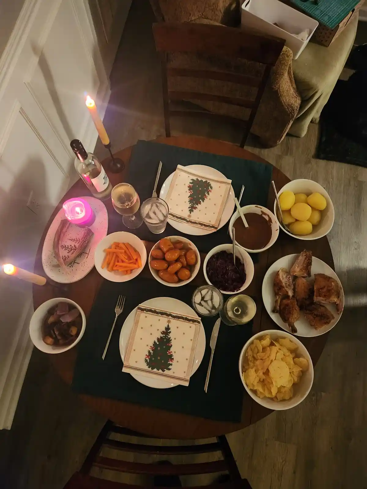 An overhead shot of a round wooden dining table set for two with festive Christmas-themed napkins, candles, wine, and…