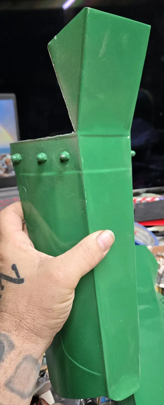 A hand holds a green, boxy, homemade-looking object with rivets, resembling a simplified, cartoonish version of a green…