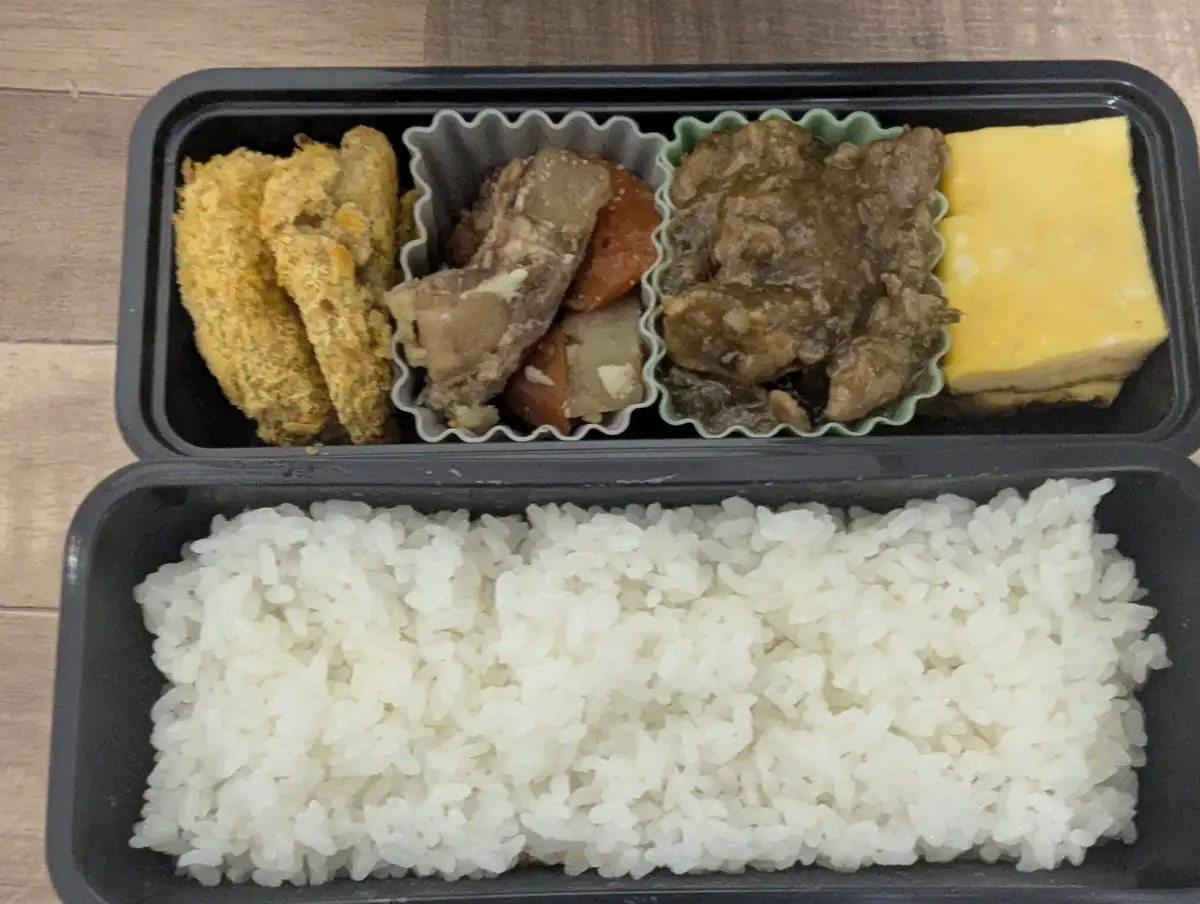 a top-down photo of a black bento box with white rice in the bottom compartment and four compartments of food above: fried…