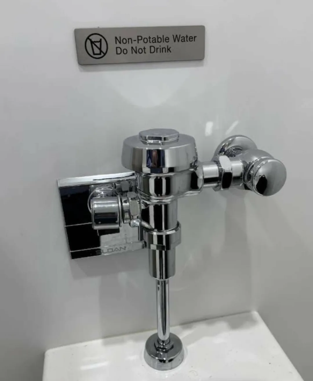 A close-up photo shows a shiny chrome flush valve for a toilet, mounted on a white ceramic fixture, with a metal sign above…