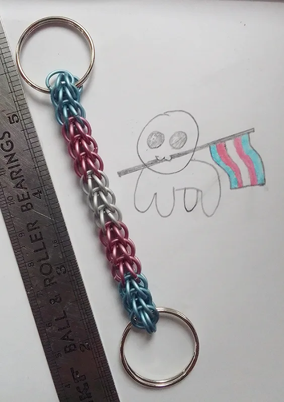 A handmade keychain made of interlinked metal rings in blue, pink, and silver sits next to a child's pencil drawing of a…