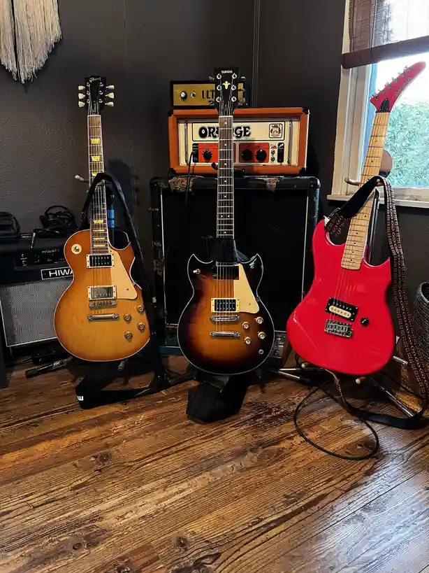 Three electric guitars — a light-wood Gibson, a sunburst Yamaha, and a bright red Kramer — stand on stands on a wooden…