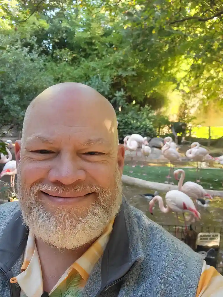 A bald man with a white beard smiles at the camera in a selfie, standing in front of a group of pink flamingos in a zoo…