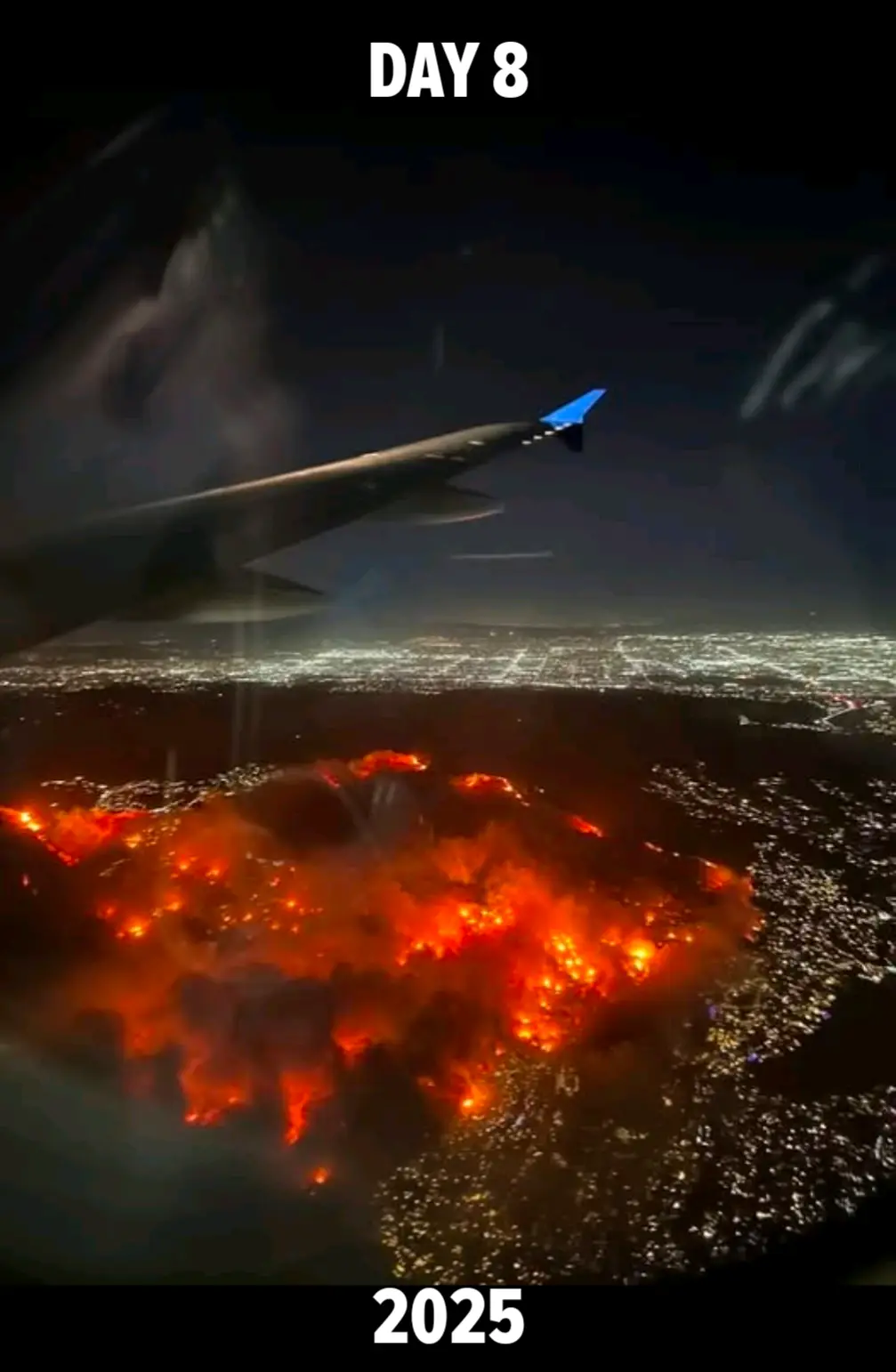 An aerial view from an airplane window shows a massive, glowing orange wildfire raging below a cityscape at night, with the…