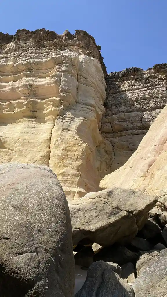 A photo of layered, sun-bleached canyon rock formations under a clear blue sky, with large boulders in the foreground. The…