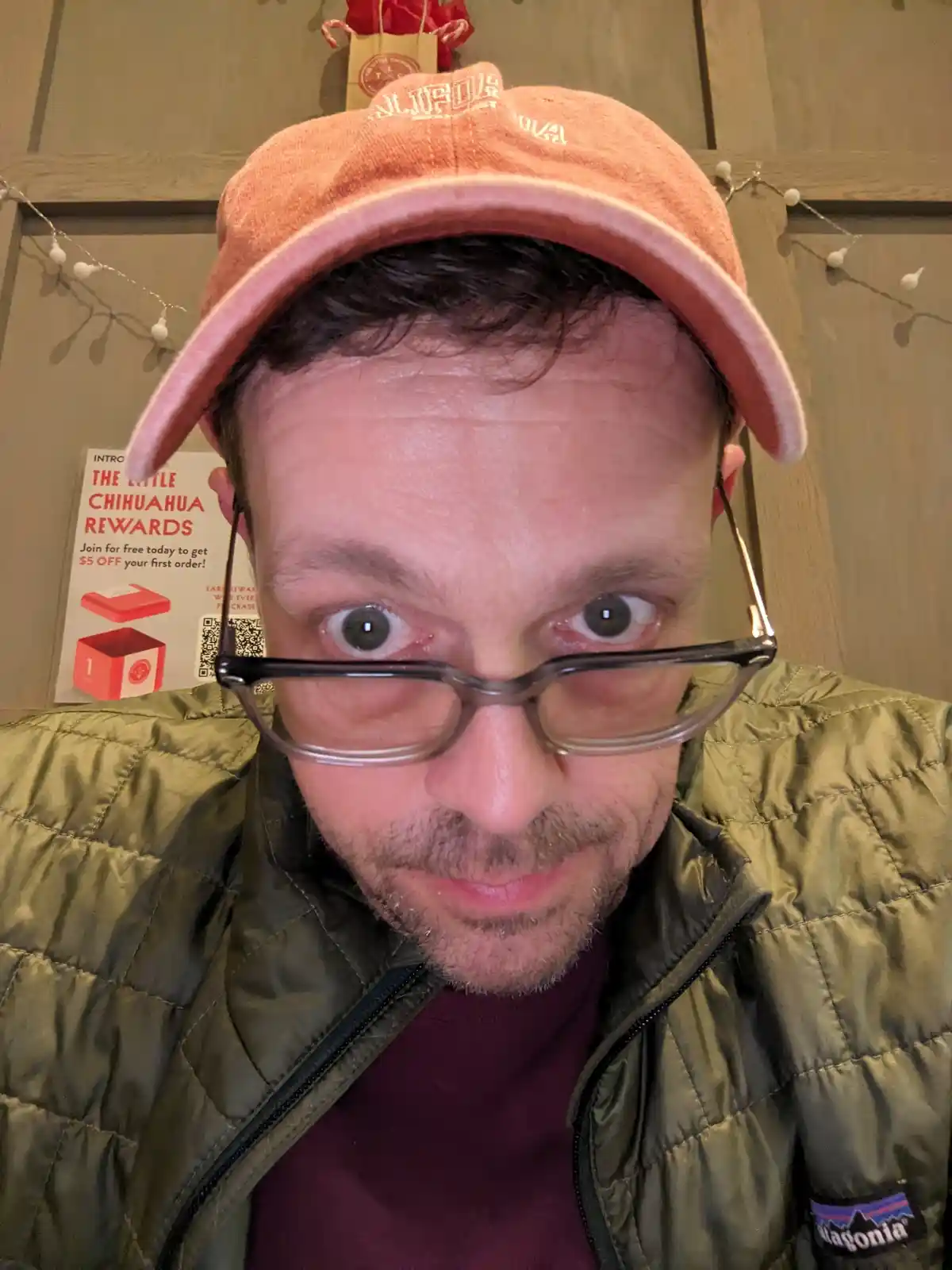 A man with wide, startled eyes and glasses looks directly at the camera, wearing a pink cap and green puffer jacket. Behind…