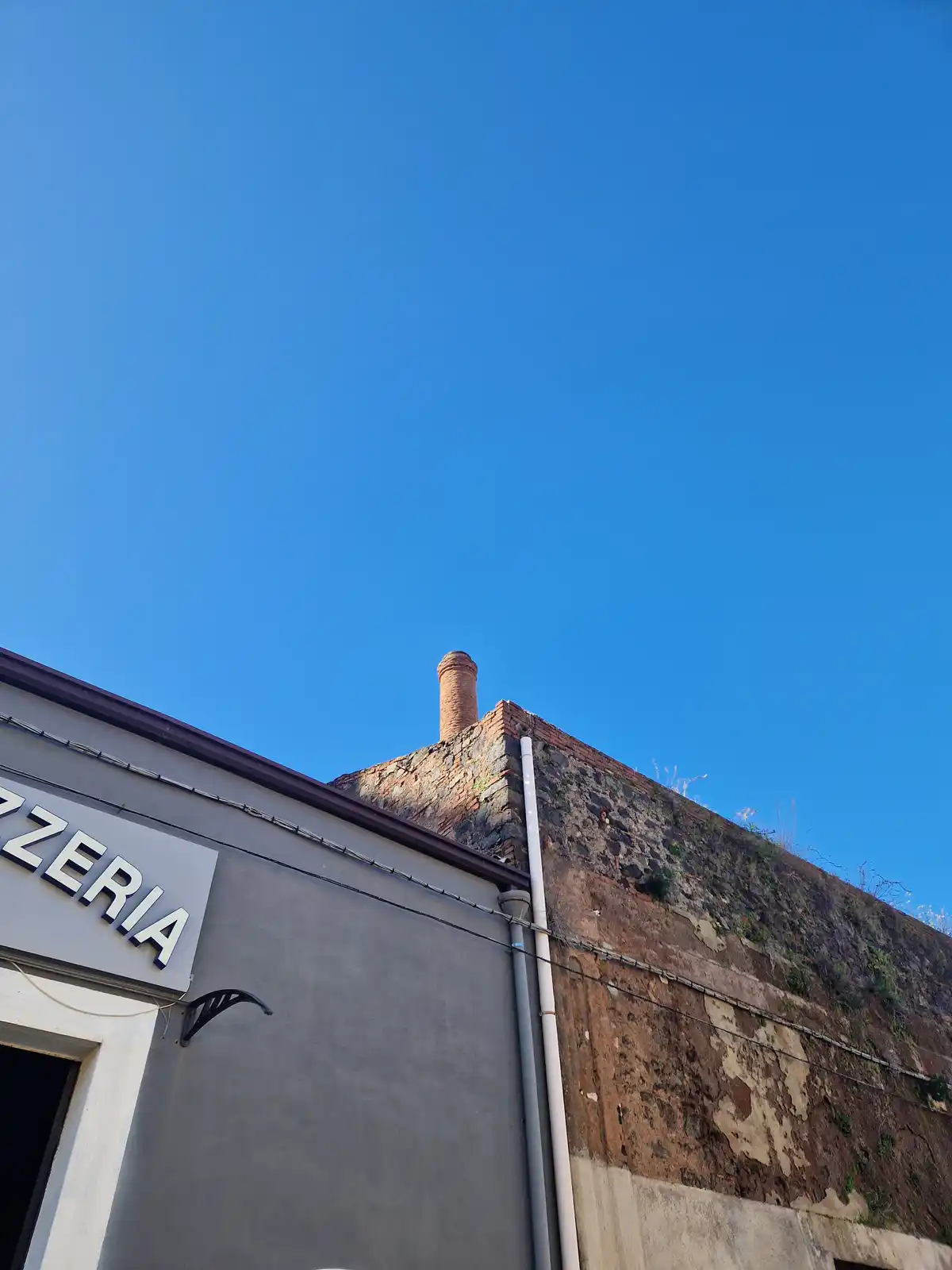 A low-angle photo shows the corner of a building with a sign reading 'PIZZERIA' and an adjacent weathered brick wall topped…