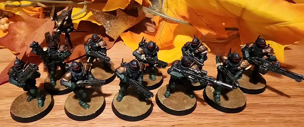 A row of painted miniature sci-fi soldiers in green and gold armor, each holding a futuristic weapon and standing on…