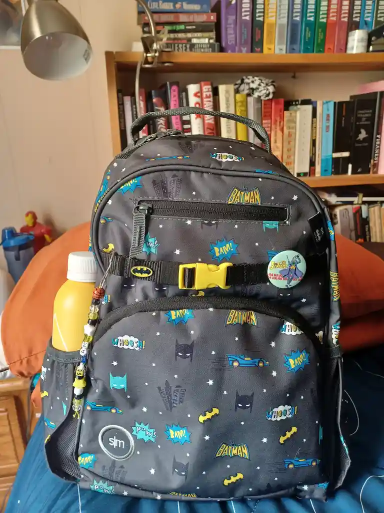 A dark gray Batman-themed backpack with comic book graphics like 'BAM!', 'WHOOOSH!', and the Batman logo is shown sitting on…