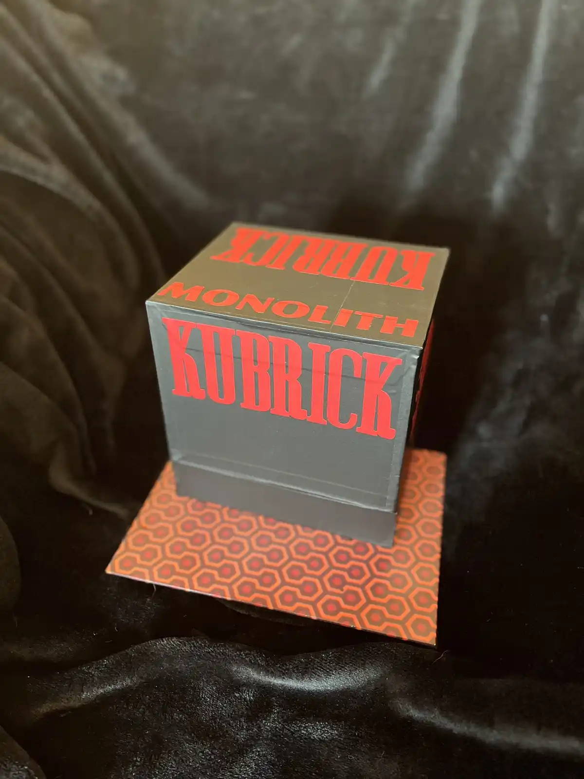 A gray cube box with red text reading 'KUBRICK MONOLITH KUBRICK' sits on an orange hexagonal-patterned coaster atop a black…