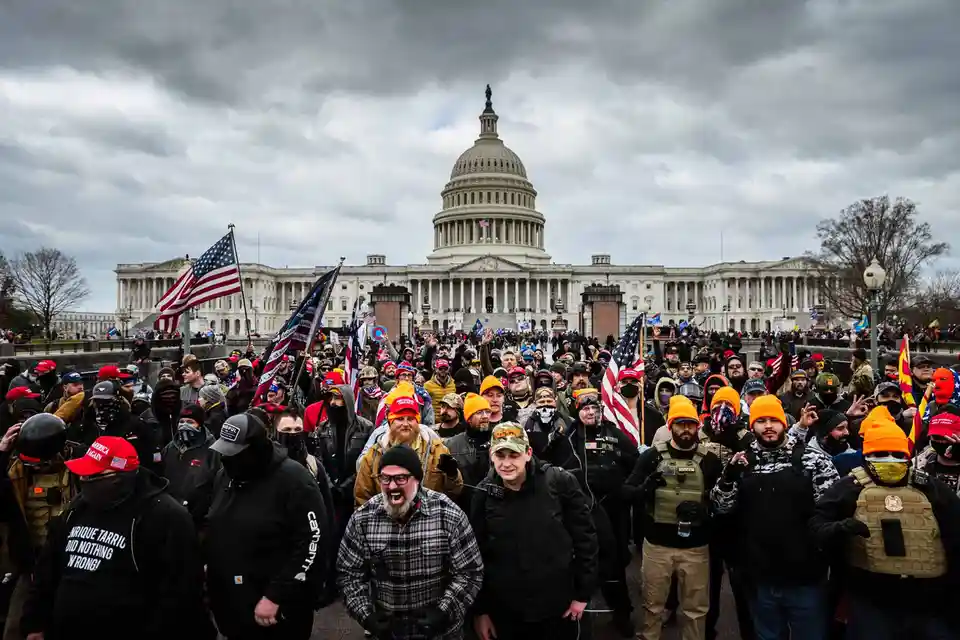A crowd of protesters wearing orange hats, red 'Make America Great Again' caps, and tactical gear stands in front of the…