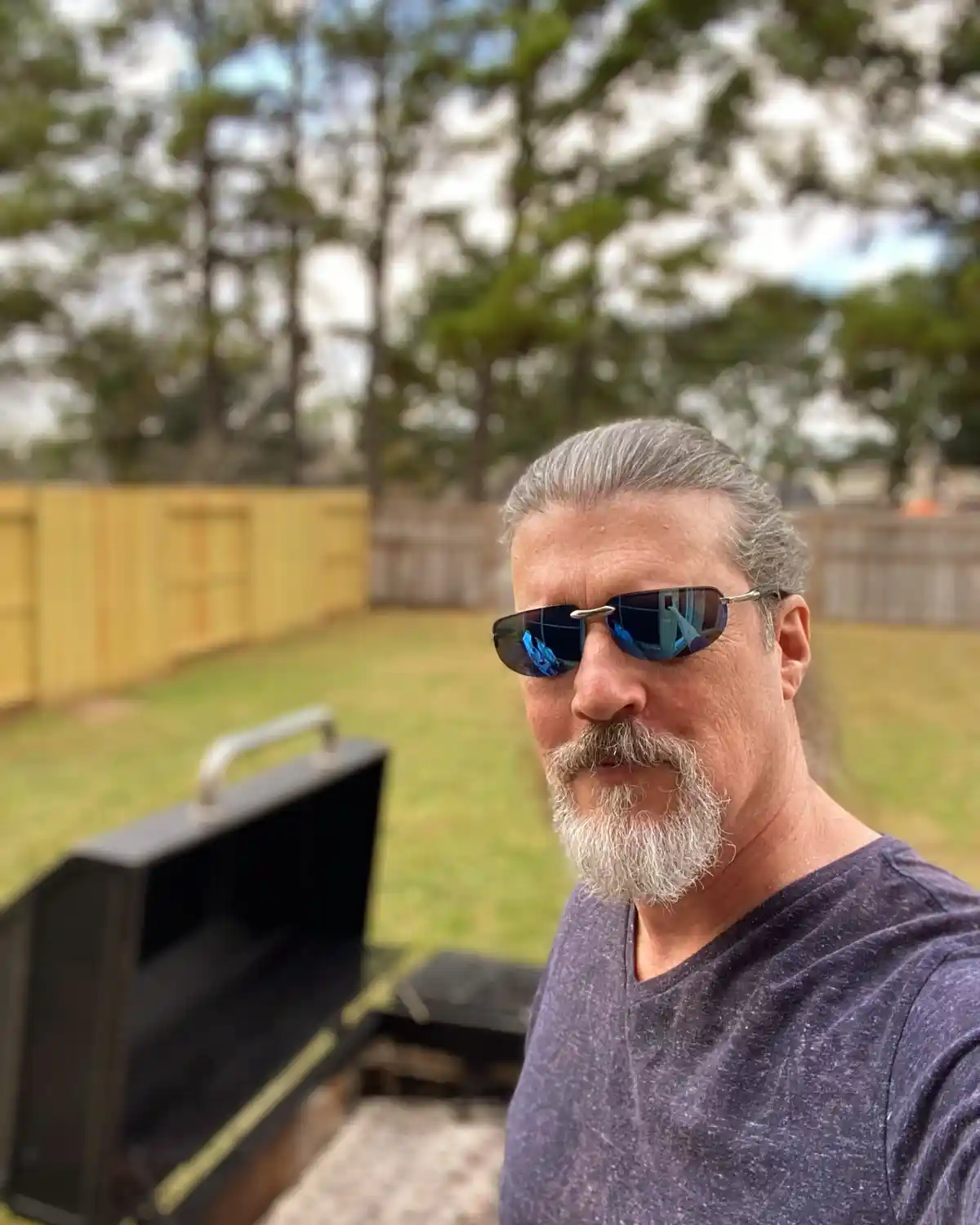 A middle-aged man with gray hair and a beard, wearing sunglasses and a purple t-shirt, takes a selfie outdoors next to an…