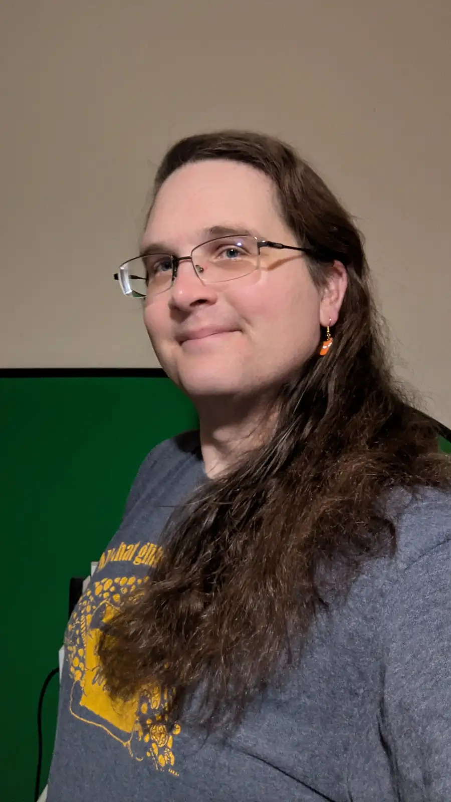 A person with long brown hair, glasses, and an orange earring smiles while wearing a gray t-shirt with a yellow graphic…